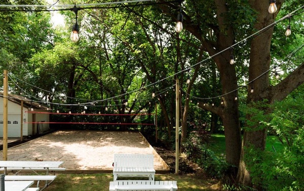 Wooded backyard gathering area with a sand volleyball court and string lights